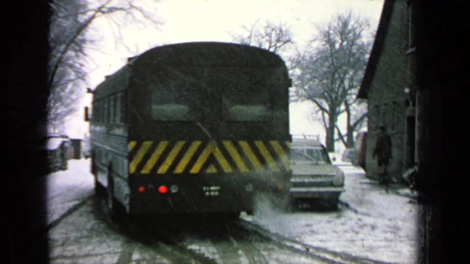 The dark bus recedes down a snow-covered base road, with a classic sedan parked nearby and a distant person near a building.