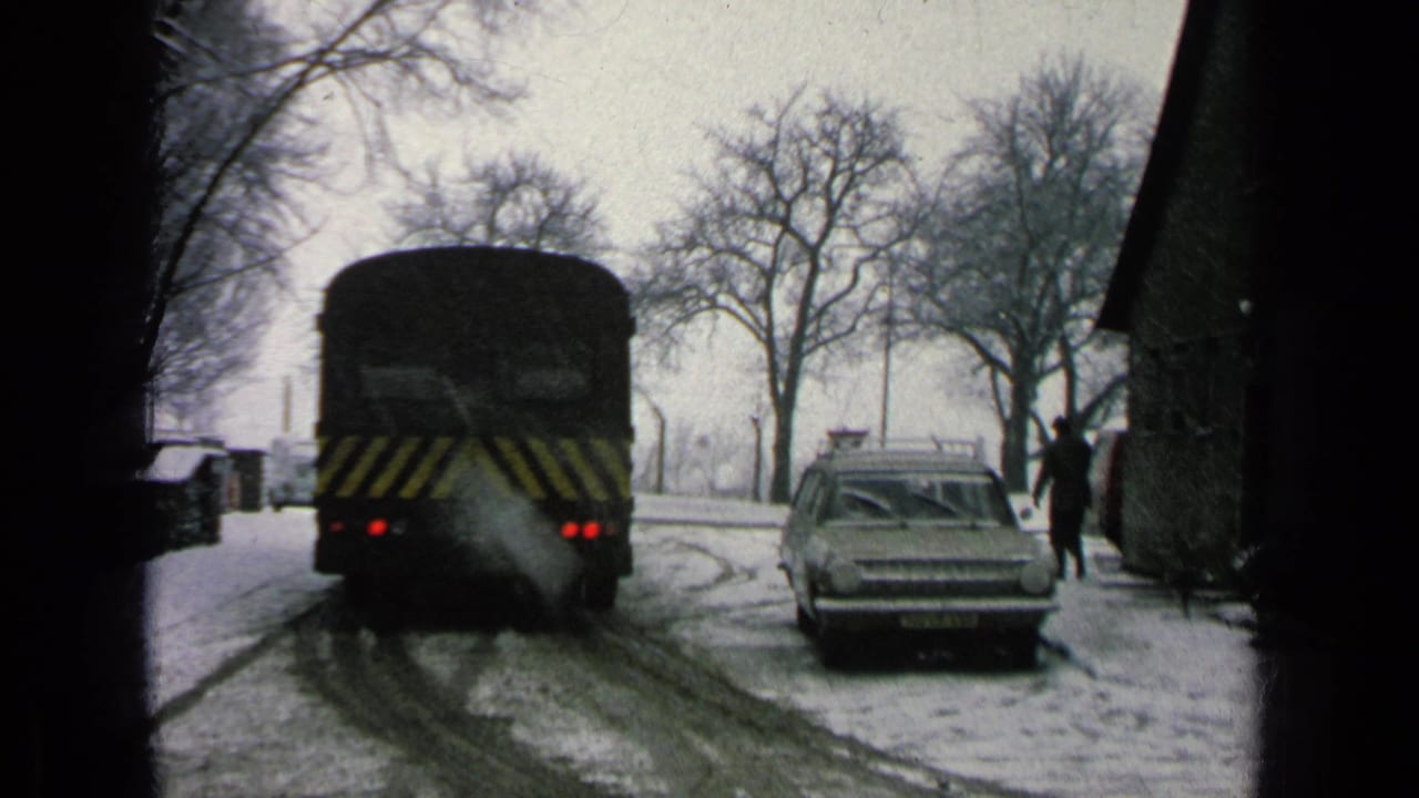 Dark bus leaving on a snowy road beside a parked station wagon and bare trees.