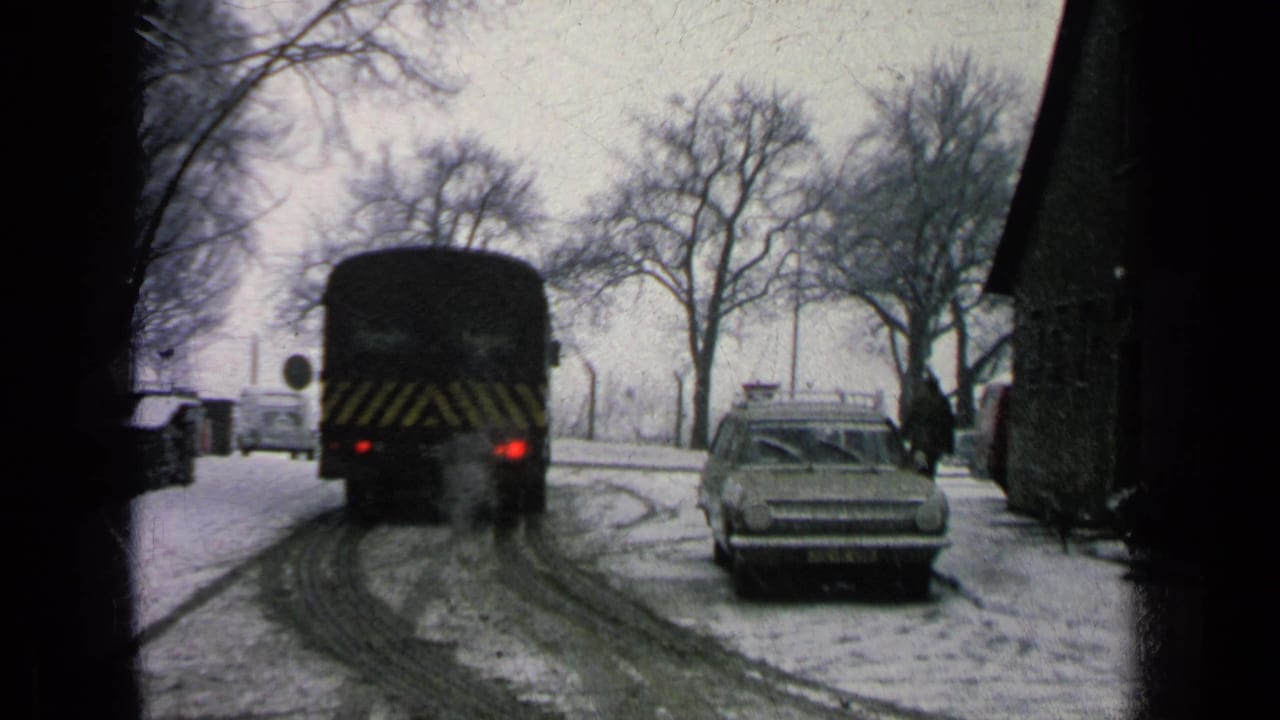 The chevron-marked bus continues away on the snow-covered road as a person stands near the building and a station wagon remains parked.