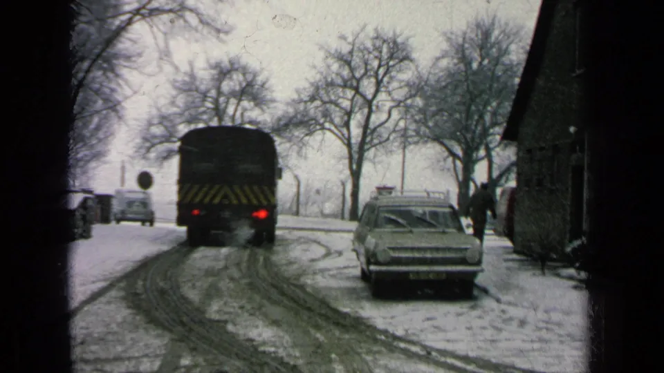 The bus moves farther from the camera on a slushy winter road, with a parked 1960s station wagon and a bundled person near the right side.
