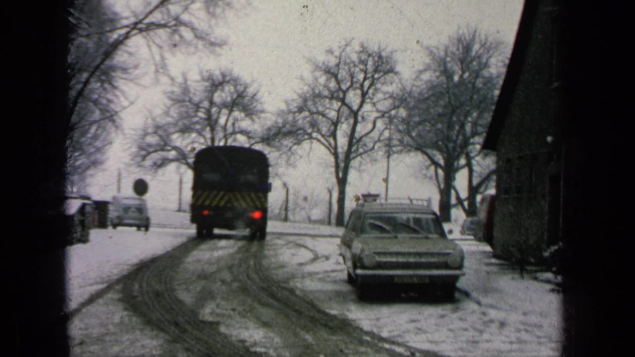 Dark bus driving away on a snowy road near a parked station wagon and bare trees.