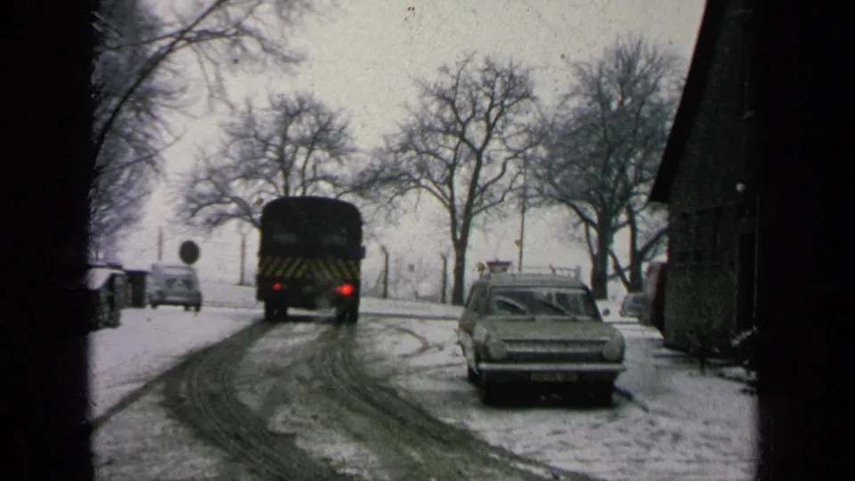 1963 8mm home-movie frame of a dark bus moving along a snow-covered base road with a station wagon nearby.