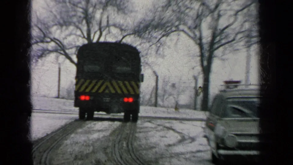 Dark bus with yellow rear striping drives through snow on a Germany military base road in 1963 footage.