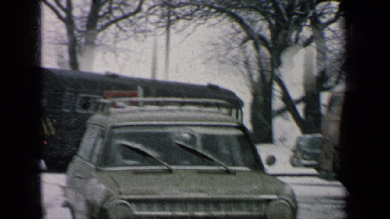 Snowy street with a parked station wagon, roof rack, bus, bare trees, and dark film-edge framing.