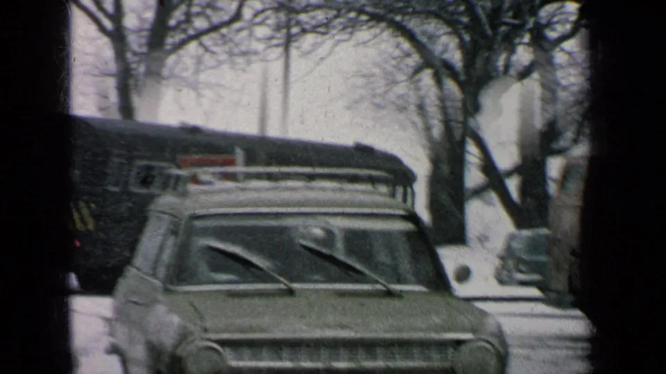 1963 8mm frame of a snowy Germany military-base street with a parked station wagon, roof rack, bus, bare trees, and soft film grain.