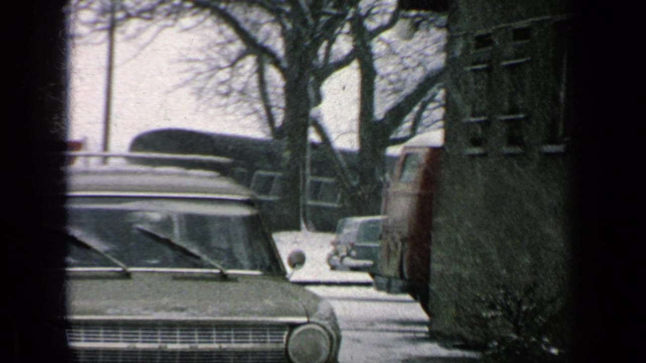 1963 8mm view of a parked station wagon on a snowy base road, with roof rack, bare trees, a signpost, and other vehicles nearby.