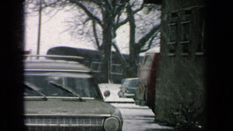 1963 8mm view of a parked station wagon on a snowy base road, with roof rack, bare trees, a signpost, and other vehicles nearby.