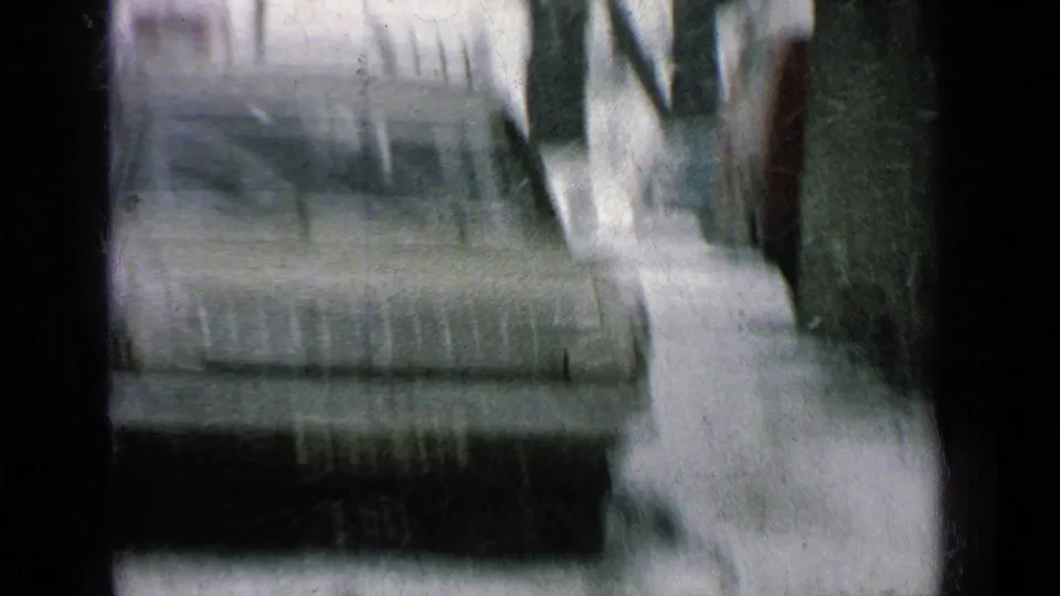 Soft, motion-blurred 8mm frame of a snowy roadside with a partial vehicle, trees, and dark vertical obstruction in the frame.