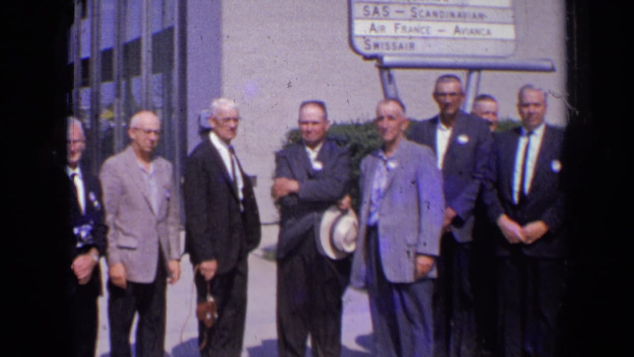 In 1963 London, adult men in suits pose outside a modern building beside a sign listing international airlines.