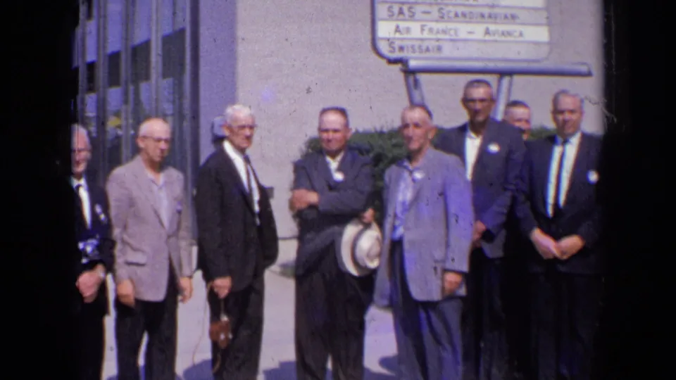 In 1963 London, adult men in suits pose outside a modern building beside a sign listing international airlines.
