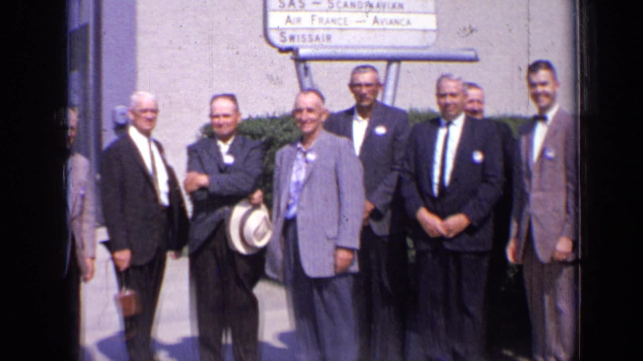 Men in 1960s formal wear stand in a row at a London airport area beneath signs for SAS, Air France, and Swissair.