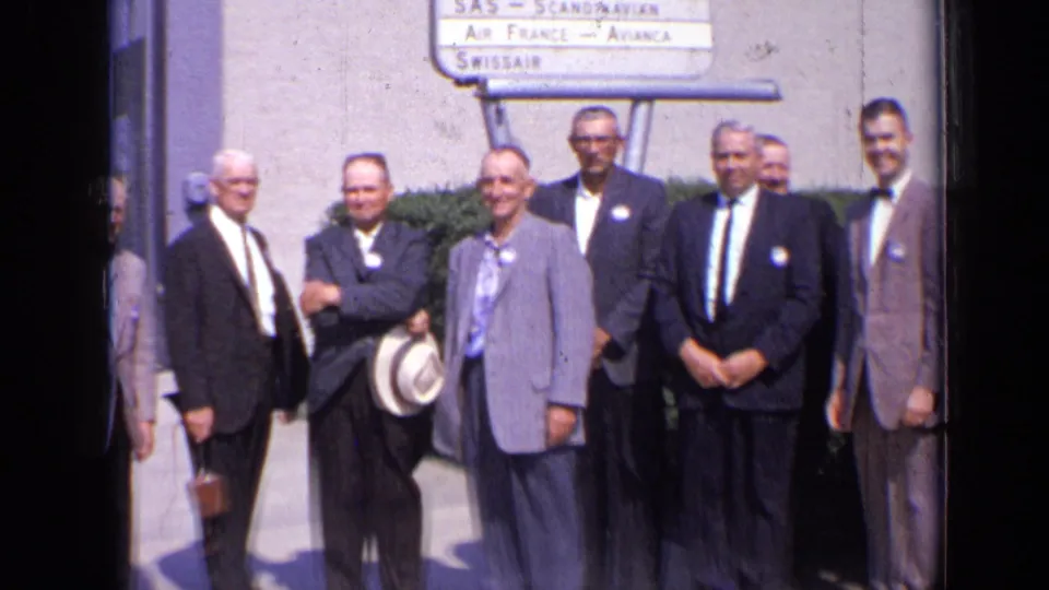 Men in 1960s formal wear stand in a row at a London airport area beneath signs for SAS, Air France, and Swissair.