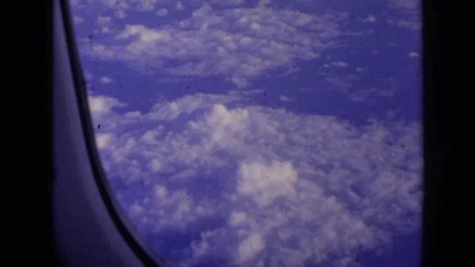 The camera looks past the aircraft window frame toward a broad layer of clouds below.