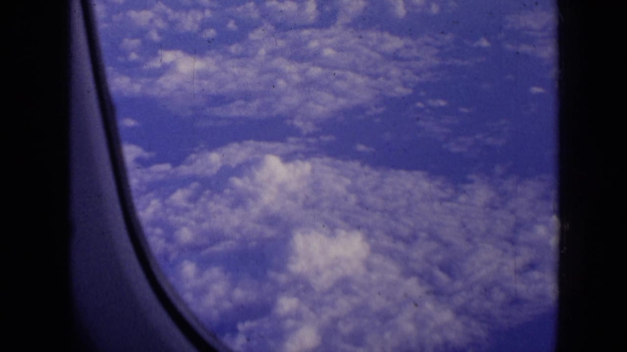 1963 8mm view of blue sky and cloud cover through a passenger airplane window, with dark cabin framing at the edge.