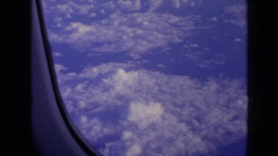1963 8mm view of blue sky and cloud cover through a passenger airplane window, with dark cabin framing at the edge.