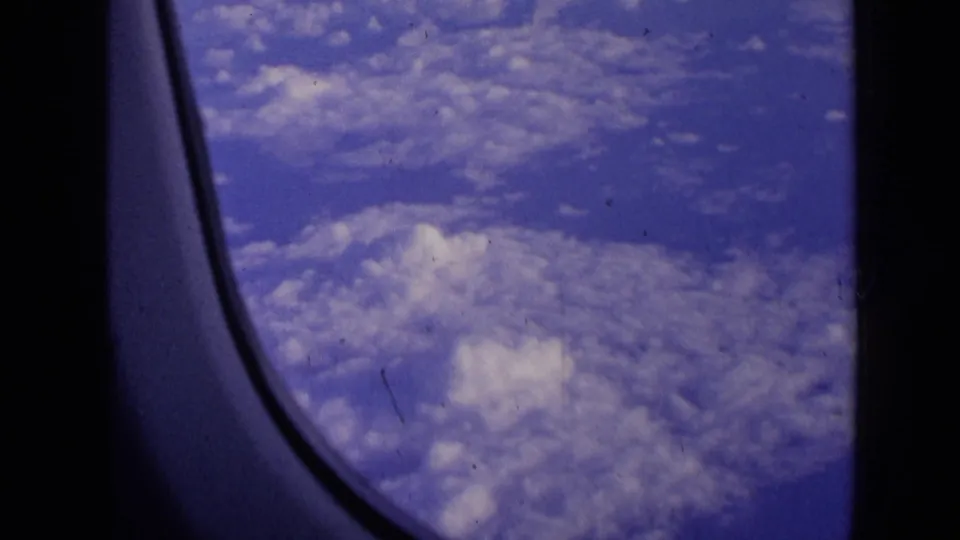 A 1963 home-movie frame looks past the airplane window curve toward layered white clouds under a blue sky.