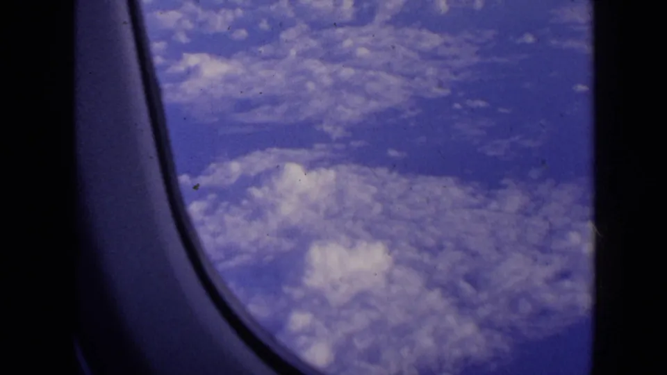 Vintage 8mm passenger-window view from flight, showing a broad white cloud layer and blue sky with visible film texture.