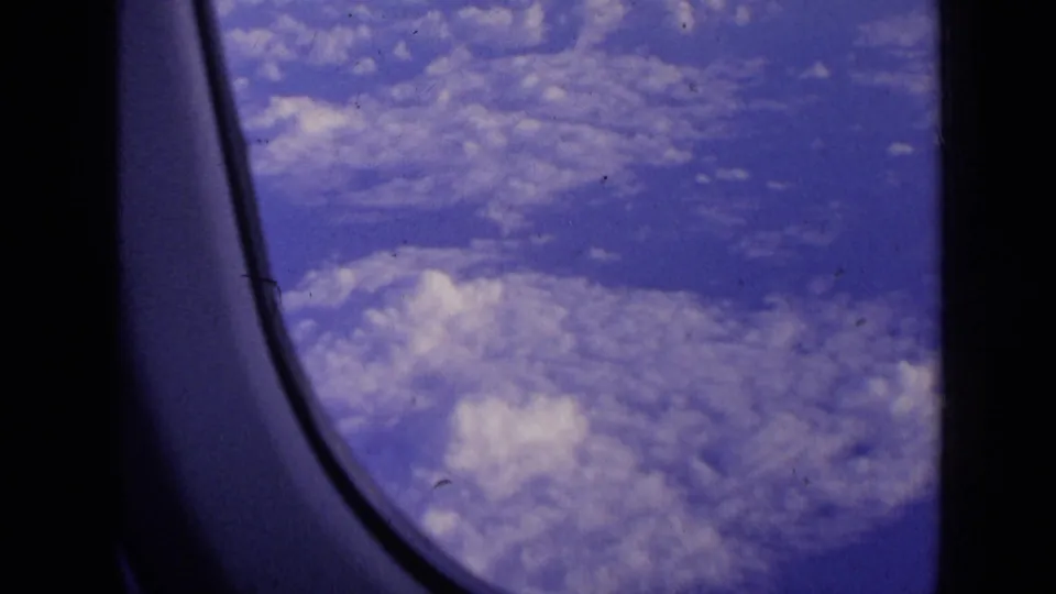 1963 8mm frame of clouds and blue sky from an airplane seat, bordered by the rounded window and dark cabin interior.