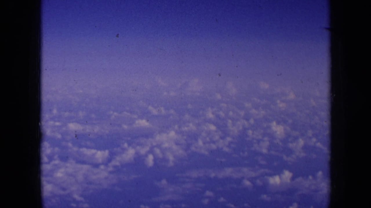 1963 8mm aerial view with scattered clouds below and a dark aircraft window divider in the foreground.