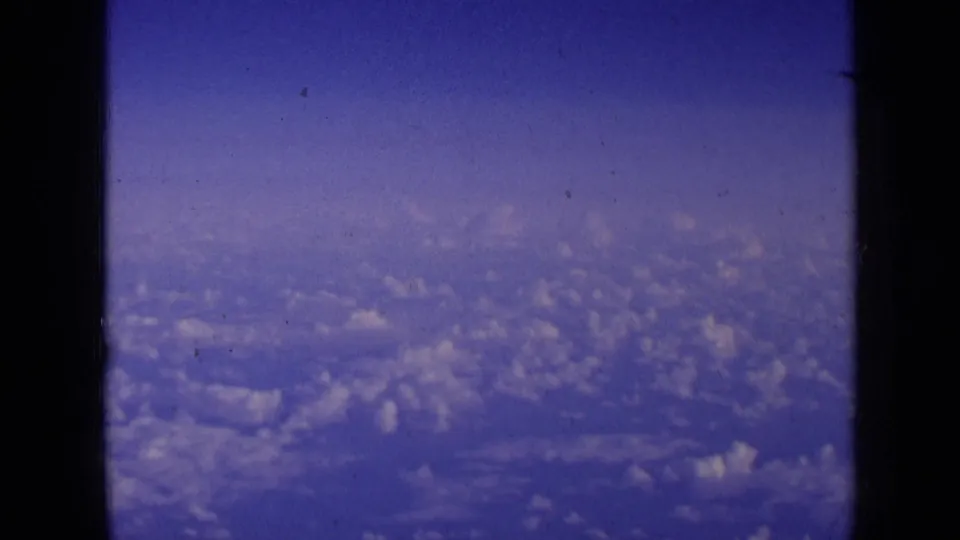 1963 8mm aerial view with scattered clouds below and a dark aircraft window divider in the foreground.