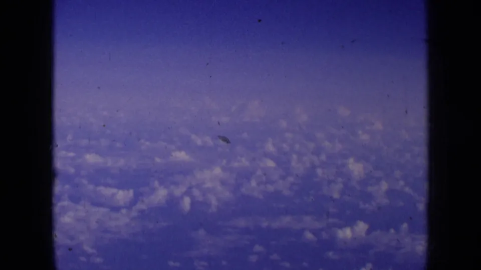 1963 8mm passenger-window view of soft blue sky and distant clouds, with dark aircraft window framing visible.