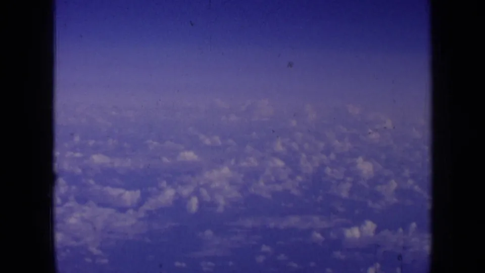 1963 8mm home-movie frame showing blue sky and cloud cover from an airplane window, with dark frame edges visible.
