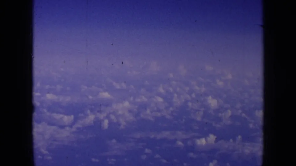 Vintage 8mm frame with clouds below and blue sky above, seen through the dark edges of an aircraft window.