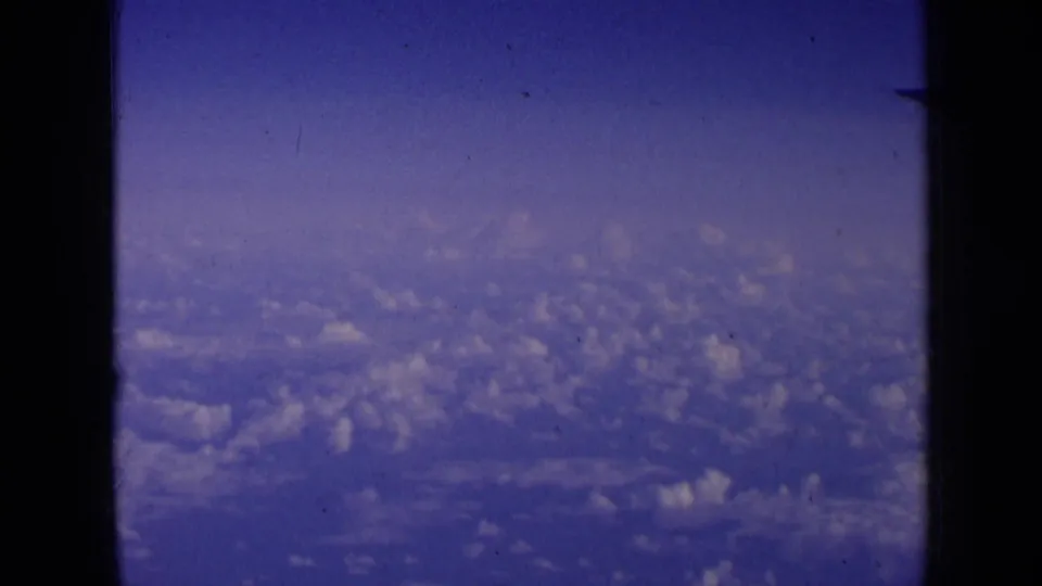 1963 home-movie aerial frame showing soft cloud formations and blue sky, with visible 8mm grain and dust marks.