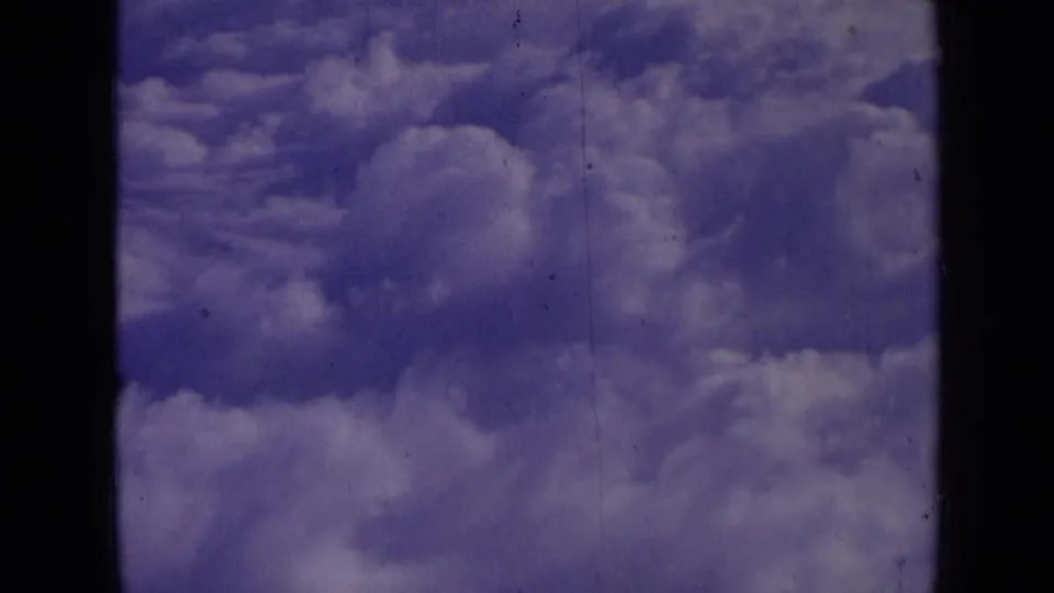Aerial cloud view from the 1963 home-movie clip, showing soft cloud forms, blue-purple color shift, and film scratches.