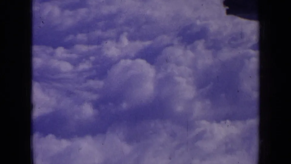 1963 8mm aerial cloud view, showing rounded white clouds, purple-blue sky, and dark frame borders from the scanned film.