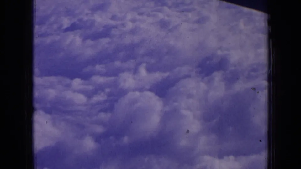 Aerial cloudscape from a passenger-window perspective, showing blue sky, cumulus forms, and aged 8mm film texture.