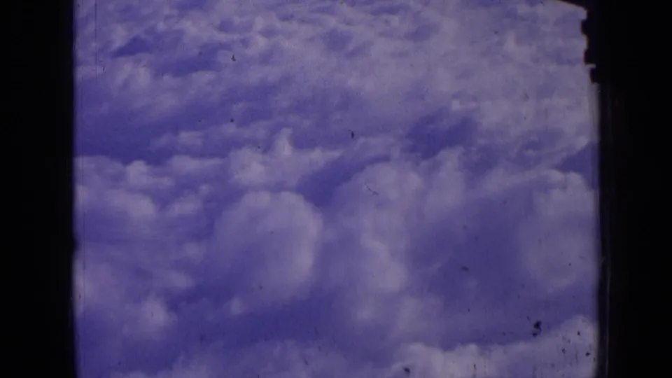 Vintage 8mm frame showing a rolling cloud blanket below the aircraft window, with grain, scratches, and muted blue tones.