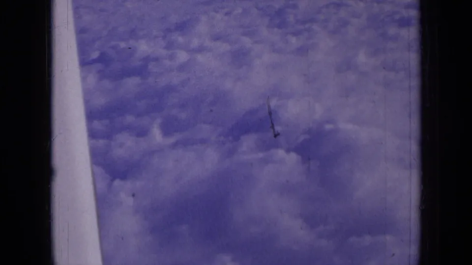 Purple-tinted 8mm view from an airplane window over cloud cover, with a pale aircraft edge visible at left.