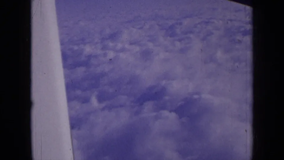 In-flight 8mm frame showing cloud cover through the airplane window, with a pale aircraft wing or strut edge visible.