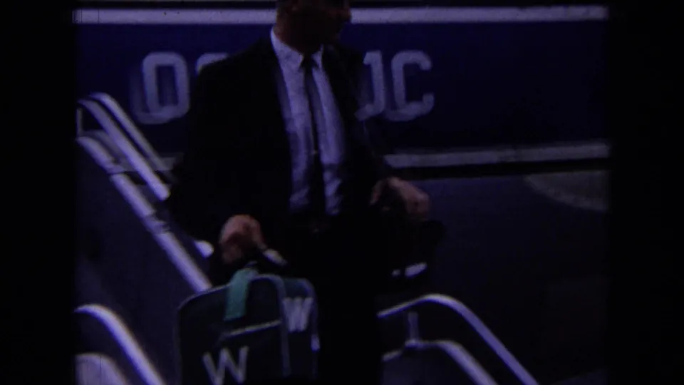 London 1963 home-movie frame of a suited passenger on aircraft stairs, holding a suitcase with a green tag near partial lettering.