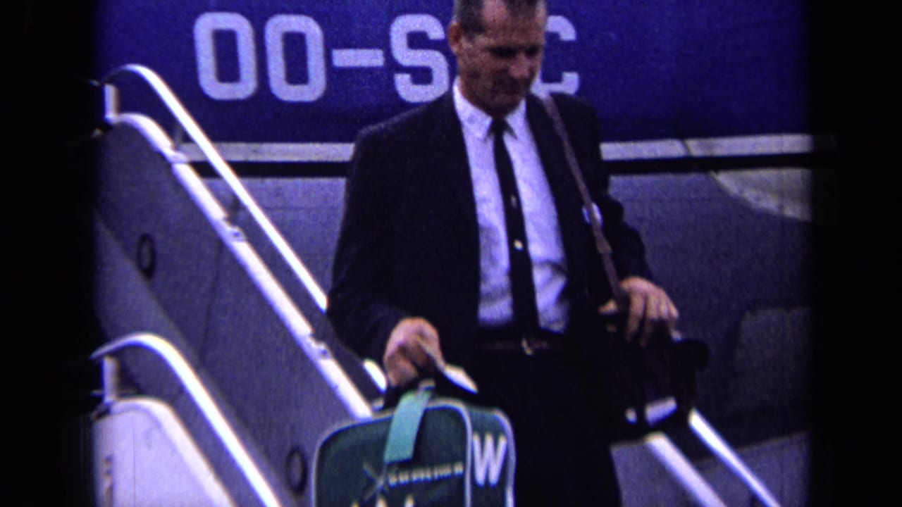 Man in a dark suit descends airplane stairs carrying luggage.