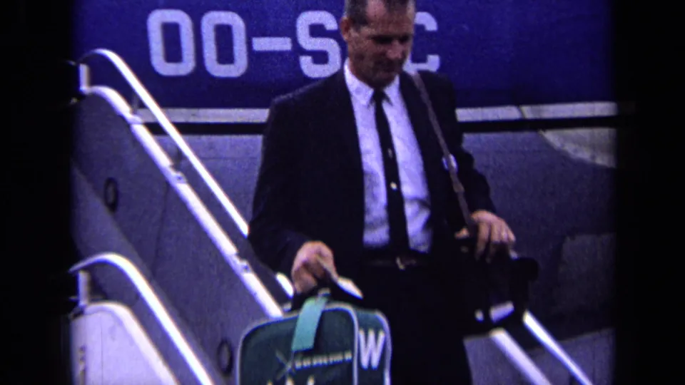 In 1963 London Airport footage, a suited man steps down aircraft stairs with a green suitcase and shoulder bag.