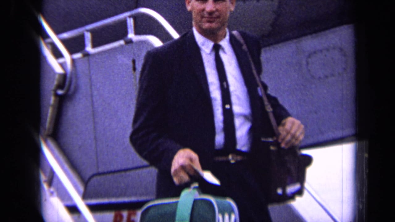The suited passenger smiles while descending aircraft stairs at the London airport location supplied in the metadata.