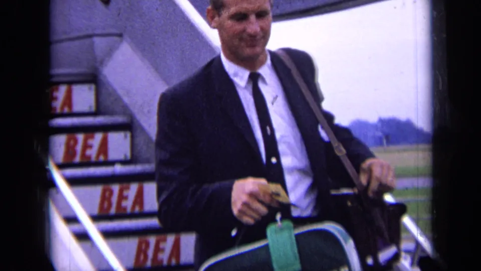 A suited traveler carries a green suitcase past aircraft stairs with repeated BEA markings visible on the risers.