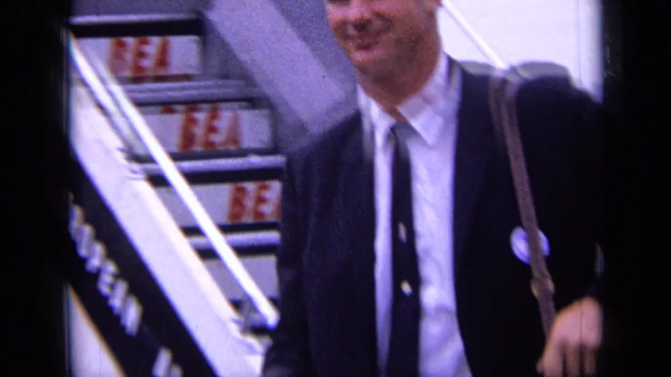 Near the bottom of the airplane stairs, the suited man with a shoulder bag passes close to the home-movie camera.