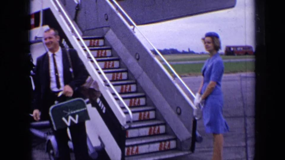 The man reaches the lower aircraft stairs with a green suitcase while the attendant in blue stands nearby at the London airport.