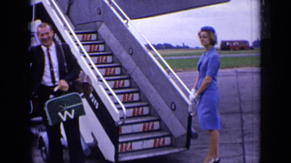 A suited passenger stands by the lower aircraft stairs as the blue-uniformed attendant waits on the tarmac in 1963 London footage.