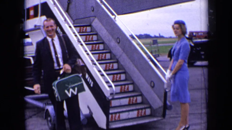 A suited passenger smiles near BEA aircraft stairs at a London airport, with a blue-uniformed attendant waiting to the side.