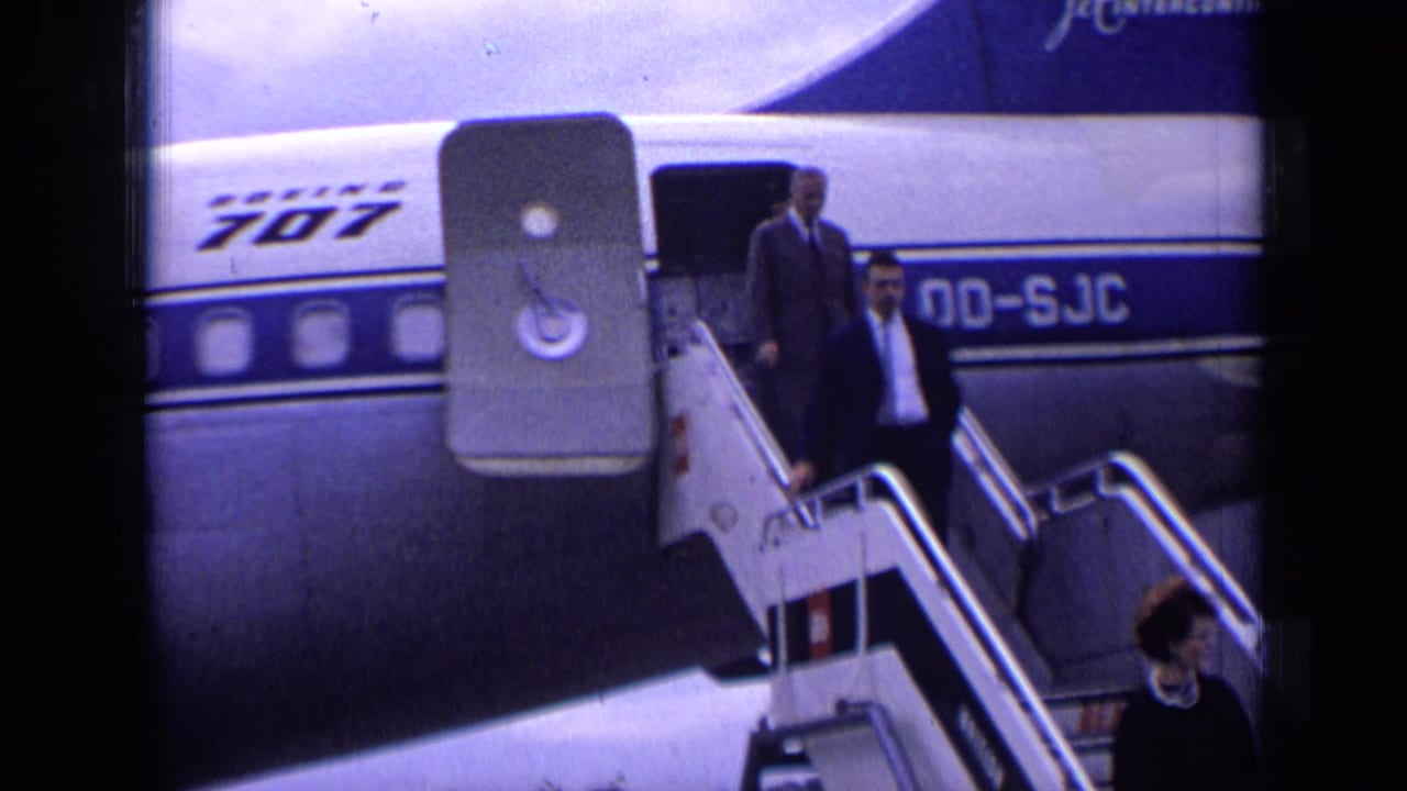 Passengers begin descending stairs from a Boeing 707 on an airport tarmac.
