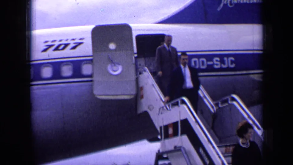 In 1963 London airport footage, formally dressed passengers descend mobile stairs from a Boeing 707 with visible fuselage markings.