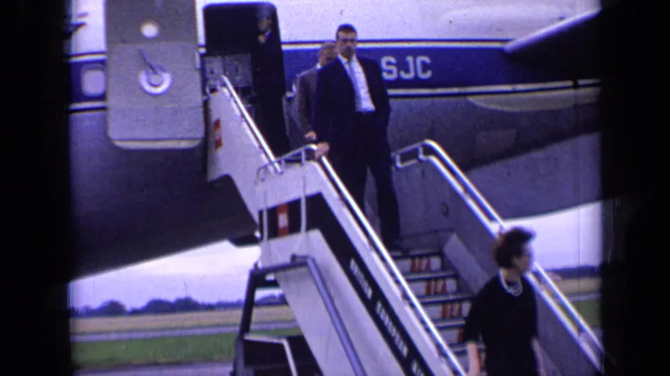 A suited passenger walks down mobile stairs from a blue-striped Boeing 707 during the London airport arrival scene.