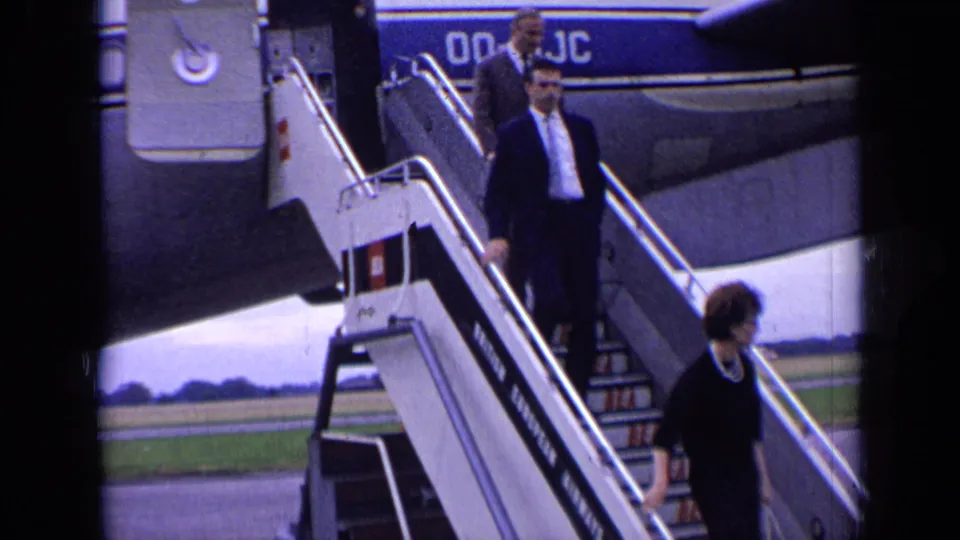 Passengers in formal 1960s dress descend the Boeing 707 stairway, with tarmac and distant grass visible behind them.