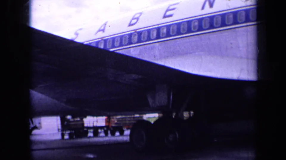 1963 London airport frame showing a partial Sabena fuselage behind a dark foreground shape, with landing gear, tarmac, and service activity below.