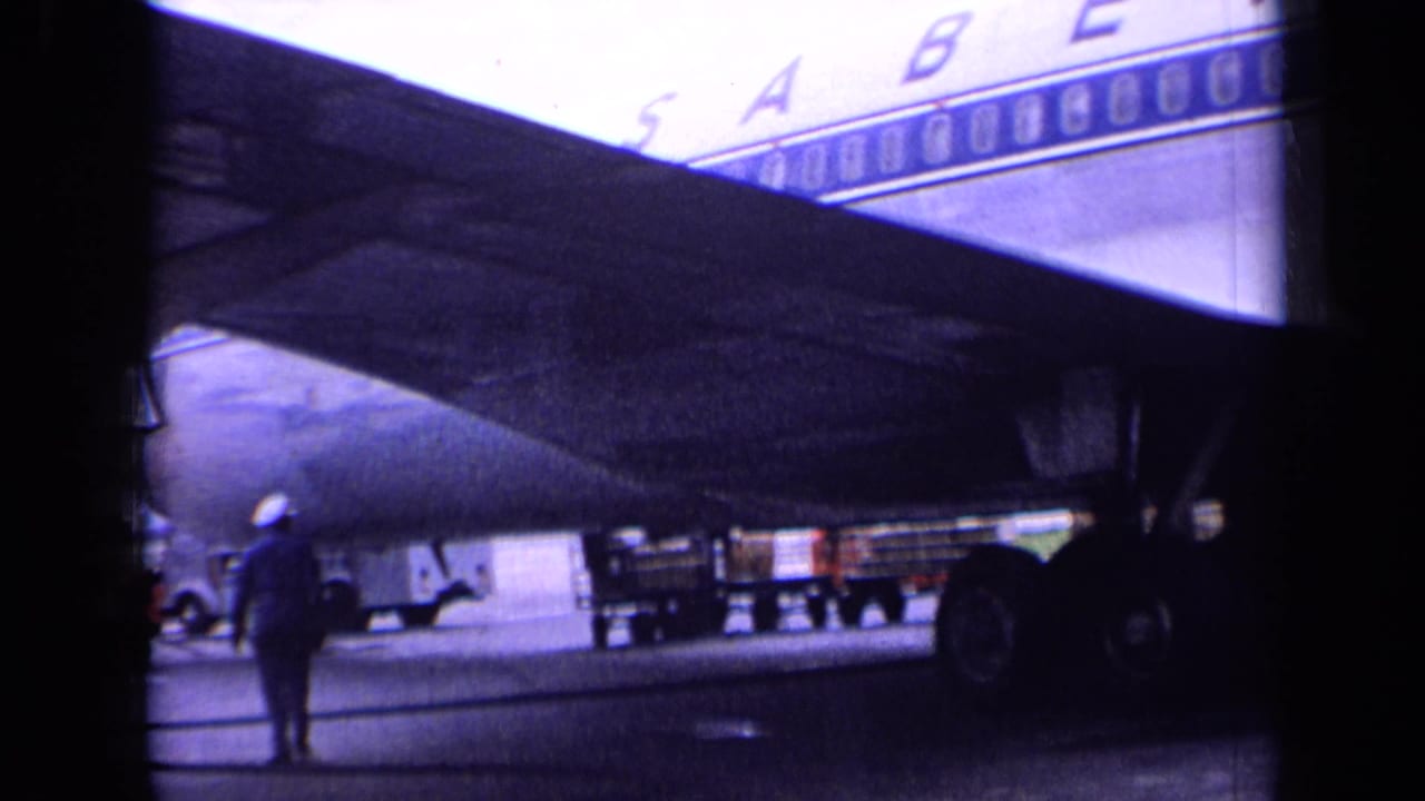 1963 London airport home-movie frame showing a Sabena airliner underwing area with ground crew and apron equipment visible.