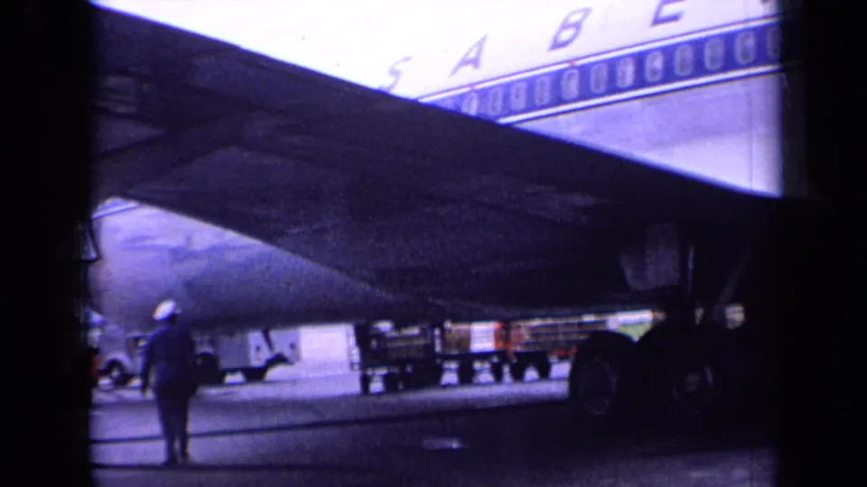 1963 London airport home-movie frame showing a Sabena airliner underwing area with ground crew and apron equipment visible.